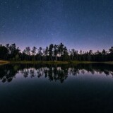 A-serene-pond-mirrors-towering-trees-and-a-starry-sky-above.-The-still-water-creates-perfect-symmetry-capturing-natures-beauty.-A-16mm-lens-with-a-long-exposure-blurs-gentle-ripples-enhancing-the-surr